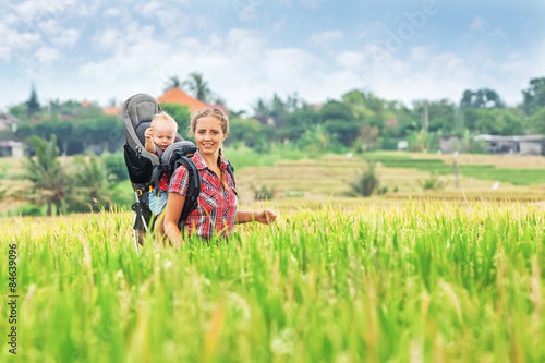 Cheerful mother with baby boy in carrying backpack walking on green rice terraces. Traveling with child, active and healthy lifestyle during family summer vacation with son on Asian tropical island