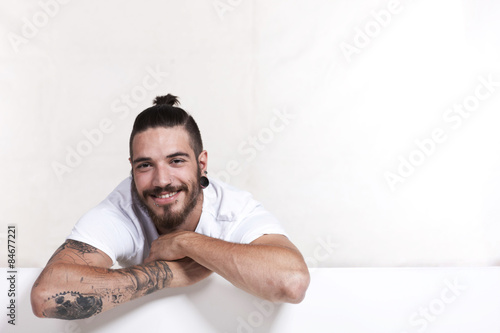 Junger Mann mit Bart und Tattoo lehnt über weißer Wand