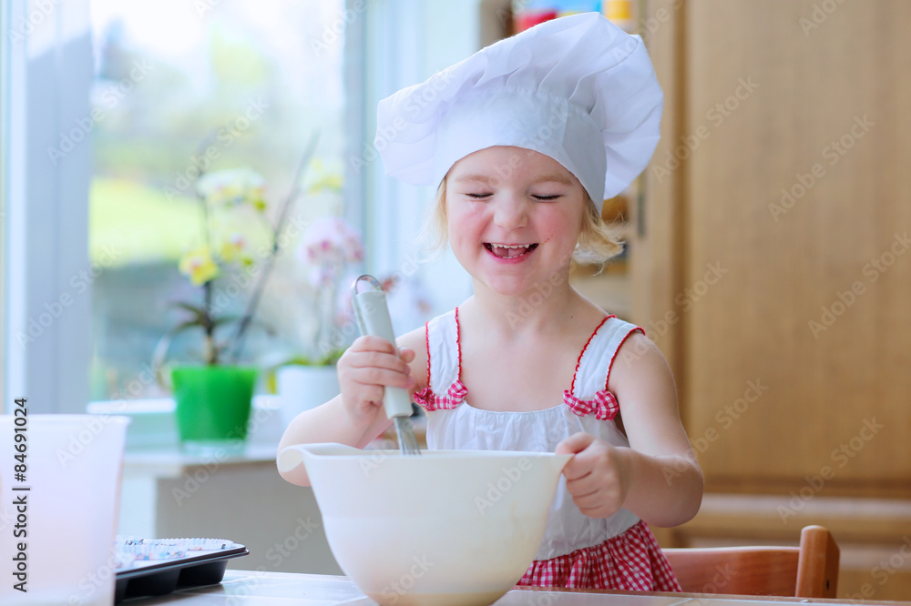 Baking with children: little happy kid, adorable toddler girl in white chef hat mixing dough ingredients in the bowl helping mother to prepare delicious pastry in the kitchen Baking with children: little happy kid, adorable toddler girl in white chef hat mixing dough ingredients in the bowl helping mother to prepare delicious pastry in the kitchen