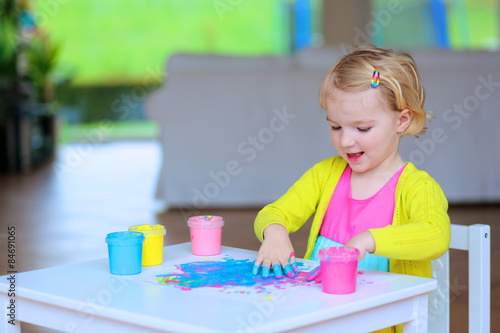 Laughing little child, blonde artistic toddler girl painting with colorful finger paints indoors at bright room at home or kindergarten. Focus on messy hands