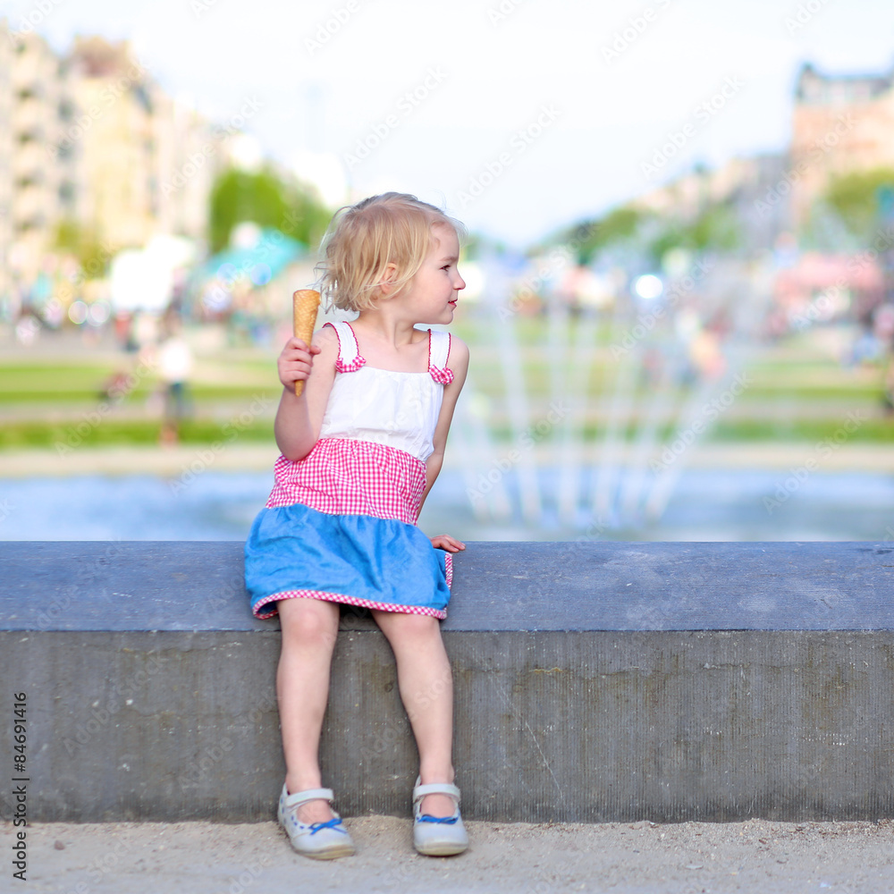 Naklejka premium Happy child enjoying summer day outdoors. Cute little toddler girl eating ice cream outdoors in city park. Beautiful blurred fountain at background.