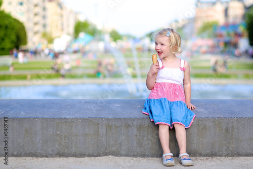 Happy child enjoying summer day outdoors. Cute little toddler girl eating ice cream outdoors in city park. Beautiful blurred fountain at background. © cromary