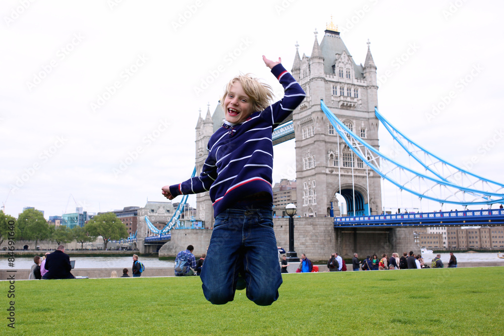Naklejka premium Chłopiec w szkole skoki na trawniku przed pięknym mostem Tower Bridge i Tamizą w słoneczny letni dzień, Londyn, Wielka Brytania. Szczęśliwy kaukaski turystyczny miły widok podczas rodzinnej wycieczki do Anglii.