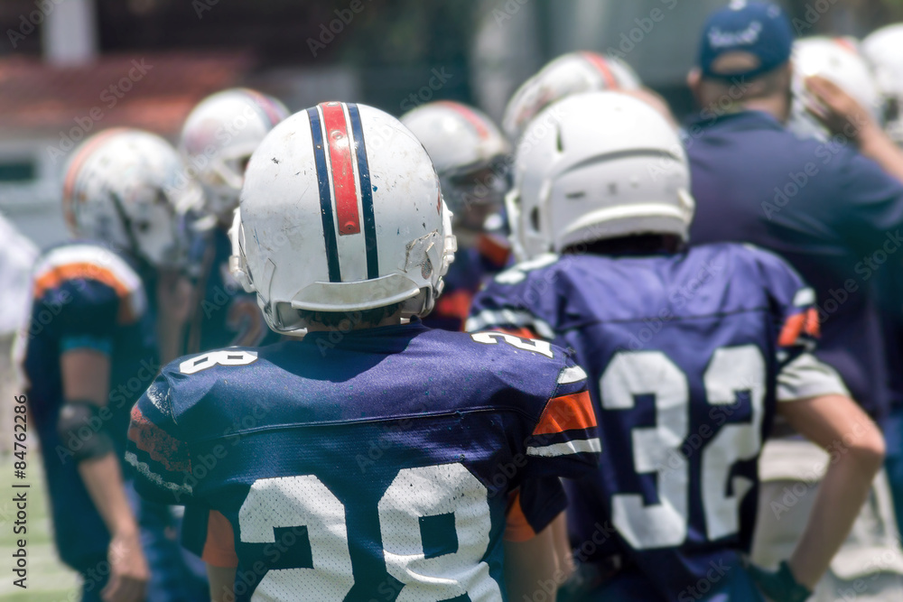 Fototapeta premium Kid standing back on a football field with white helmet and blue jersey watching other kids play 