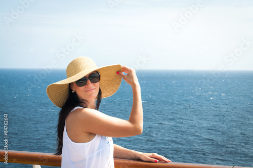 Woman wearing a floppy straw hat and a white dress standing next to the railing on a cruise ship
