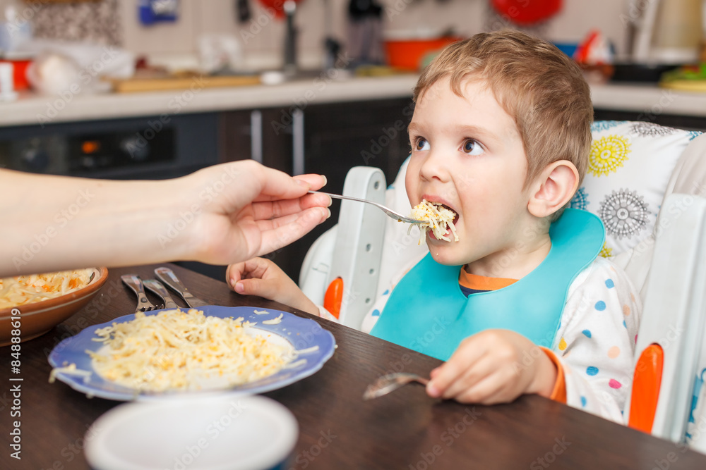 Mother feeding hungry boy in the highchair indoors Mother feeding hungry boy in the highchair indoors