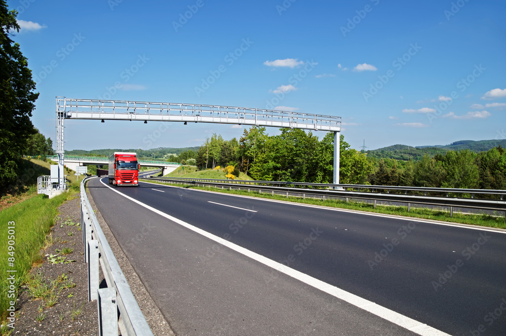 Red truck arriving toward electronic toll gate over a highway in a wooded landscape