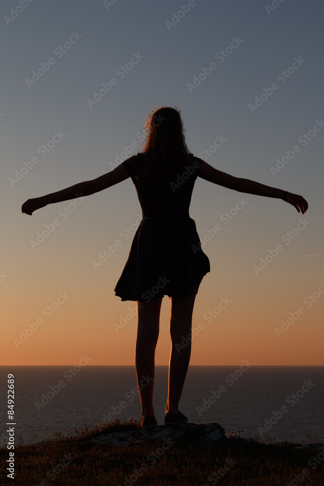 Teenage girl standing silhoutted on the cliffs at sunset looking out to sea with her arms spread wide.