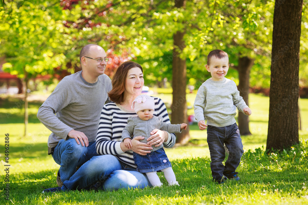Fototapeta premium Portrait of mother, father, daughter and son, happy family of four, sitting on the meadow in the park outside on a spring summer bright day