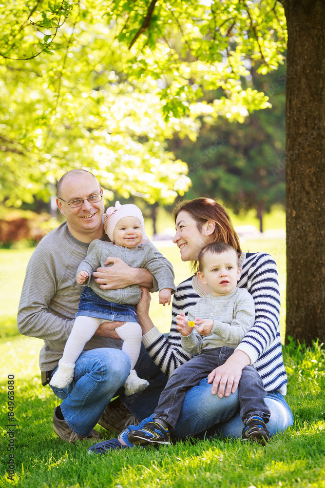 Portrait of mother, father, daughter and son, happy family of four, sitting on the meadow in the park outside on a spring summer bright day