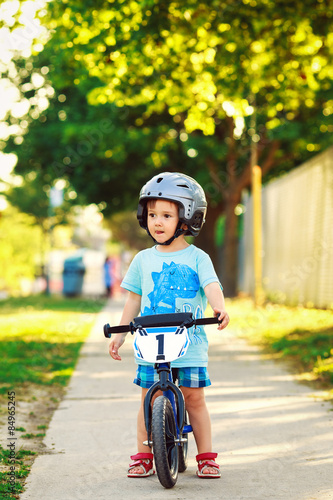 Fototapeta Naklejka Na Ścianę i Meble -  Portrait of a little boy toddler riding a bicycle in helmet on the road outside outdoors on a bright sunny spring summer day, seasonal child activity concept, healthy lifestyle