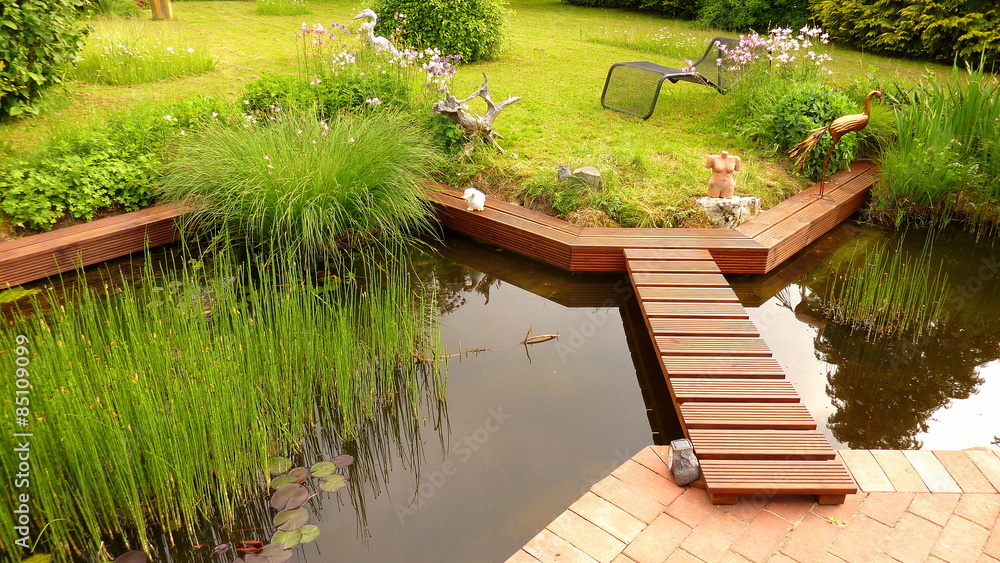 Gartenteich im Grünen umrandet mit Holzdielen und kleiner Holzbrücke
