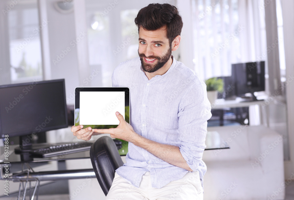 Portrait of young casual businessman sitting at office and holding white screen digital tablet in his hands while looking back and smiling.