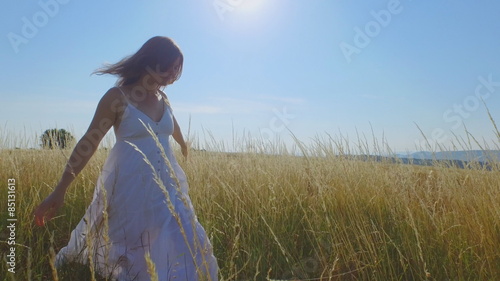 Beautiful young woman with a long white dress walking through long grass and gently touching the stems.