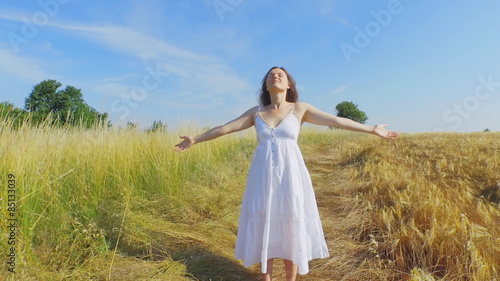Beautiful young woman with a long white dress standing in a field of long grass lifting her arms up with joy of being surrounded  by natural beauty.