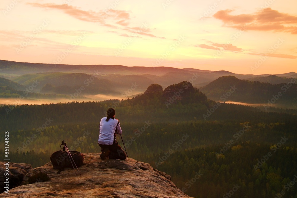 Fototapeta premium Professional photographer in white jacket takes photos with camera on tripod on rocky peak. Dreamy fogy landscape, spring orange pink misty sunrise in a beautiful valley below