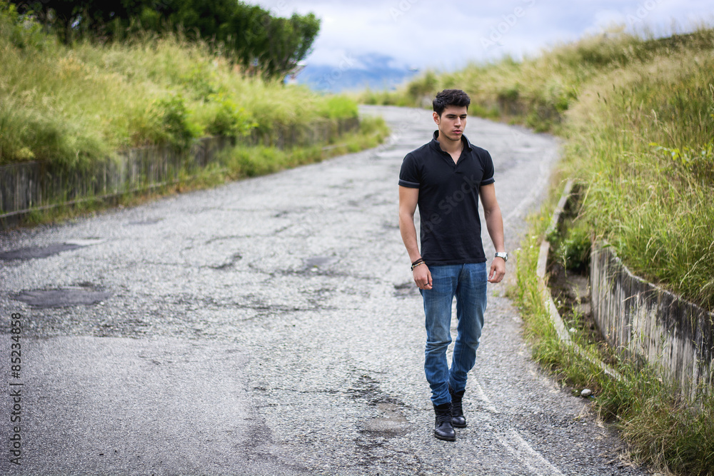Fototapeta premium Young man in jeans and black t-shirt walking along rural road