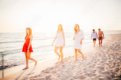 A group of five friends walking on the beach at sunset. Two young men and three young women in small groups, they leave behind the sun and walking on the sand in a day of rest.