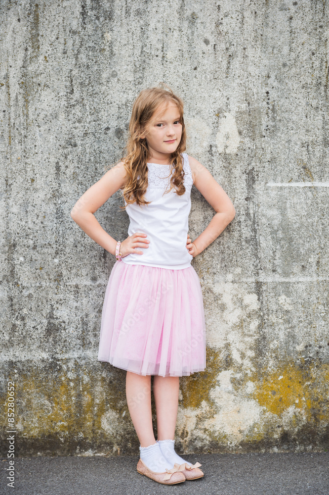 Close up portrait of a cute little girl of 7 years old, standing against grey wall in a city