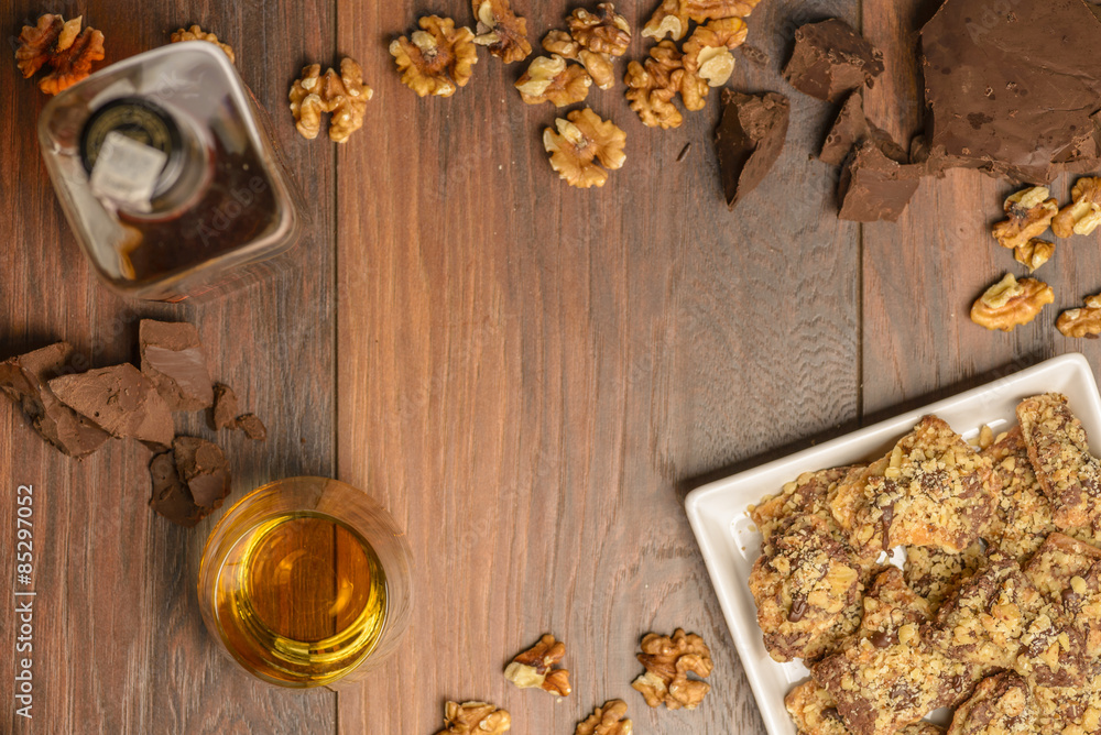 Overhead view of homemade chocolate walnut cookies with ingredients and a bottle and glass of scotch whisky on a dark rustic wood table. Open space in center for logo or text.