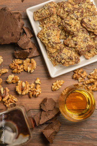 Overhead view of homemade chocolate walnut cookies with ingredients and a bottle and glass of scotch whisky on a dark rustic table.