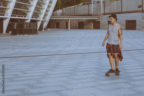 cool young and handsome caucasian brunette hipster skater guy wearing a hat posing smiling and having fun outside while skating with his skateboard during amazing summer day in the city