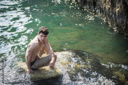 Young shirtless athletic man sitting on rock by sea