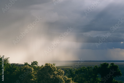 An intense rain storm approaches, its rain clouds intercepted by the sunrays of the sunset, making for a fantastic view. Registered from a mountain top, in the midst of the jungle, in Cambodia.