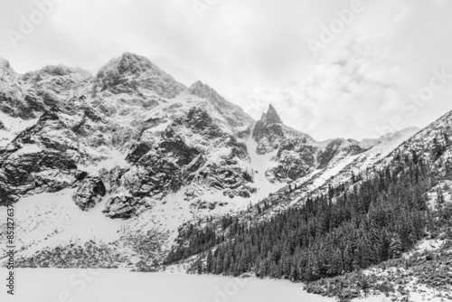 Black and white landscape of a frozen snow covered alpine lake and mountain peaks on a cloudy winter day.