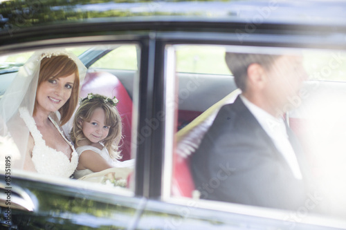 Bride sitting in car with little bridesmaid and driver before wedding photo