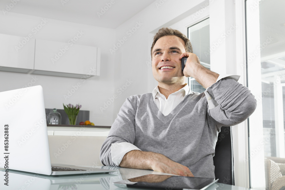 Smiling man sitting in kitchen with laptop and digital tablet telephoning with smartphone Smiling man sitting in kitchen with laptop and digital tablet telephoning with smartphone