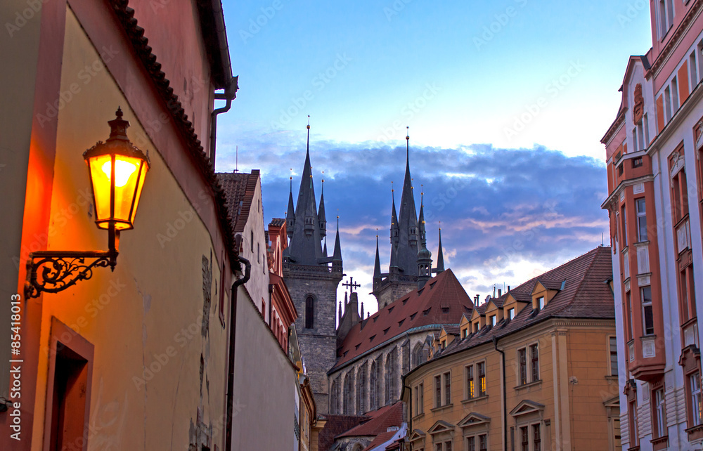 Prague by twilight - shape of the steeples of the gothic cathedral against evening sky and brightly yellow lit old street lamp on the foreground.