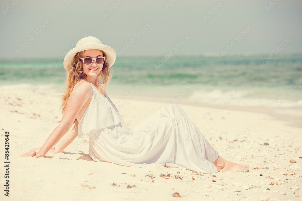 Young woman in long white dress and hat relaxing on tropical beach having great summer time. Vacation, lifestyle, travel, concept