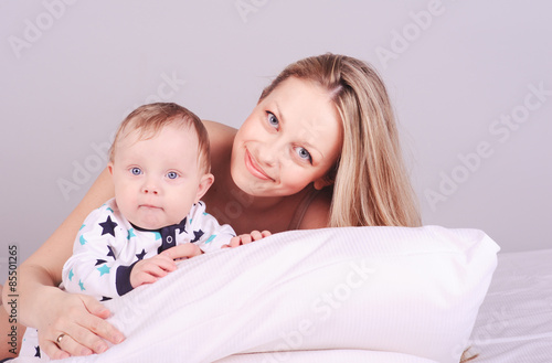Smiling young woman lying in bed with baby boy. Happy mother holding newborn child over gray. Motherhood