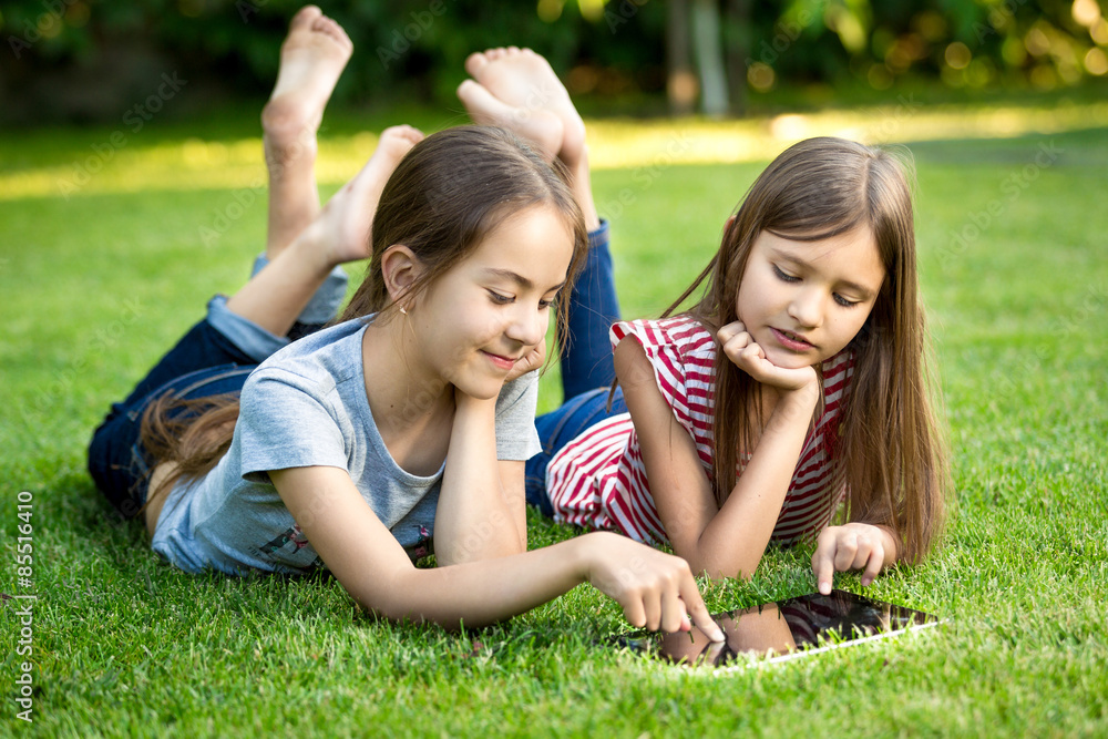 Fototapeta premium Two sisters lying on grass outdoors and playing on digital table