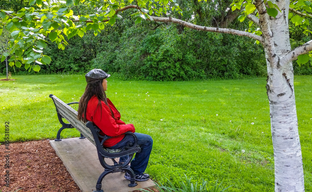 Fototapeta premium man with long black hair, black cap,red jacket and jeans sitting on bench relaxing in a park staring ahead