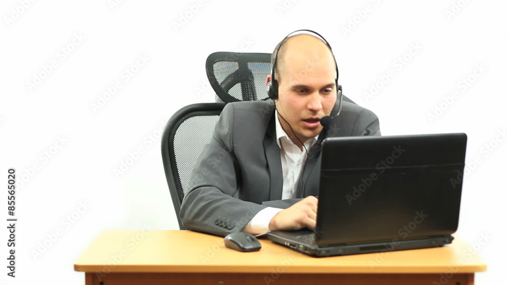 Guy from technical support sitting behind a black laptop, dressed in a grey suit and white shirt without a tie, using headphones and a microphone. Guy from technical support sitting behind a black laptop, dressed in a grey suit and white shirt without a tie, using headphones and a microphone.