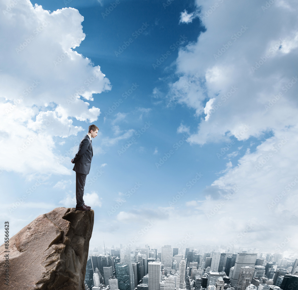Young businessman standing on edge of rock mountain and looking at the city Young businessman standing on edge of rock mountain and looking at the city