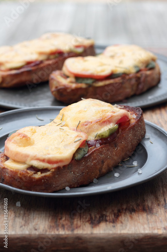 Freshly grilled slice of bread with pickles, tomatoes, ham and melted cheese.