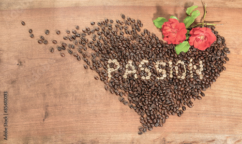 Chicchi di caffè con rose rosse, disposti in forma di cuore su un piano di legno grezzo.Scritta con chicchi di caffè crudo.