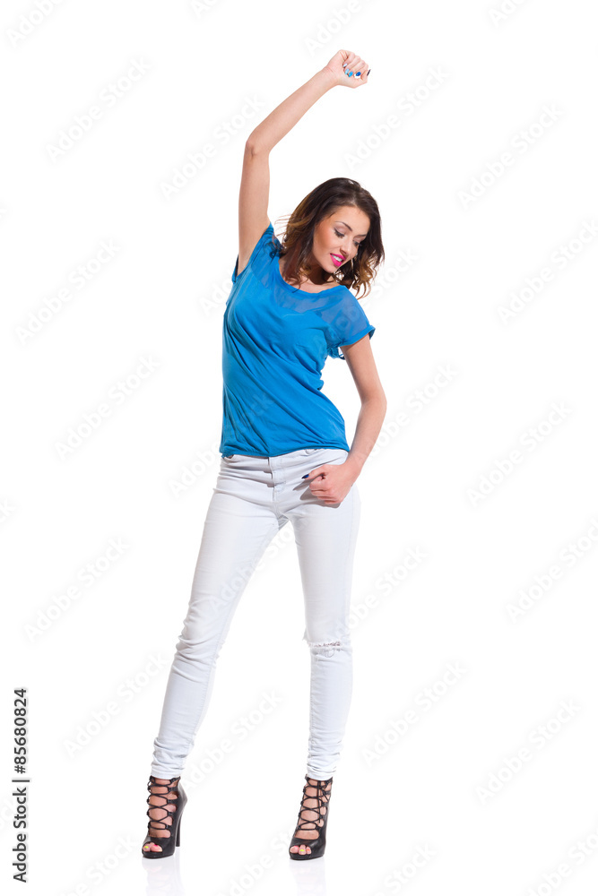 Smiling young woman in blue top, jeans and high heels posing with arm raised and looking away. Full length studio shot Isolated on white. Smiling young woman in blue top, jeans and high heels posing with arm raised and looking away. Full length studio shot Isolated on white.