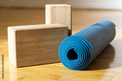Yoga Mat and Blocks - Yoga Accessories in Studio. A blue yoga mat with two wooden yoga blocks sit on a wooden yoga studio floor before class.