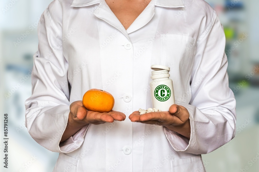 Young doctor holding mandarin and bottle of pills with vitamin C and compare them Young doctor holding mandarin and bottle of pills with vitamin C and compare them