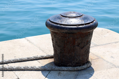 Rusty mooring bollard on a dock with nautical rope. Bright blue sea in the background.