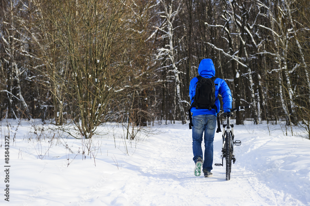 Young extreme cyclist with black backpack, in jeans and blue jacket walking with mountain bicycle on snowy track near forest in winter sunny cold day 