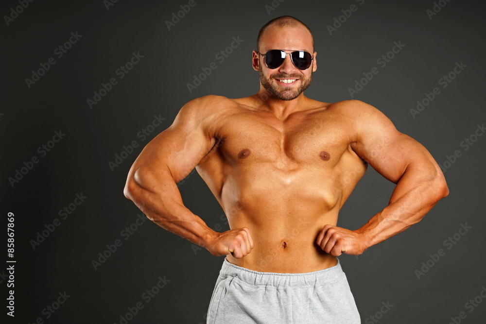 Muscular man in sunglasses shows his muscular body on gray background