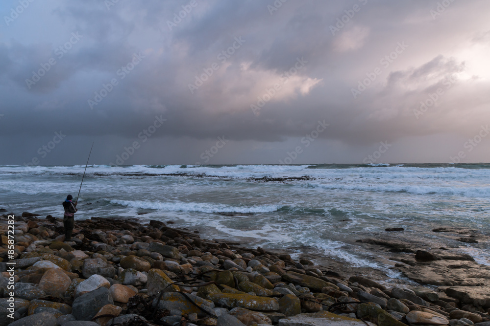 Lone fisherman on an isolated, rocky beach under a dramatic stormy sky on the South African coast