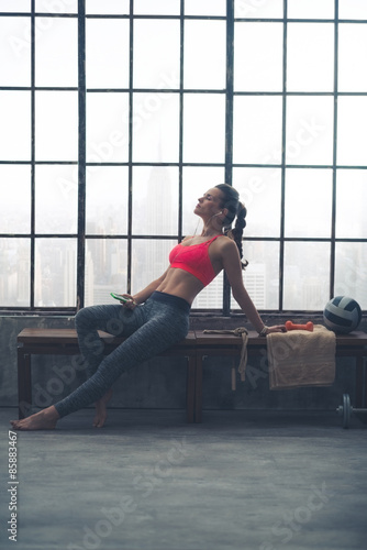Fit, muscular woman relaxing on bench in loft gym with earbuds