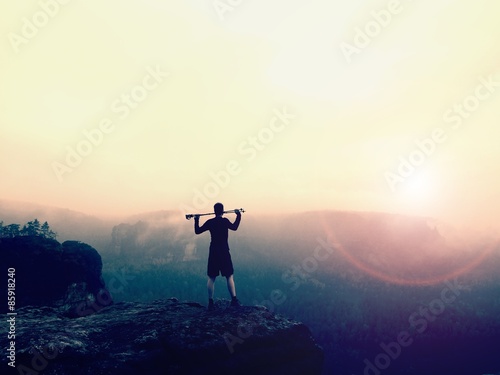 Tourist with poles in hands is standing on rock and watching into orange misty landscape. National rock empire park, melancholic autumn morning.