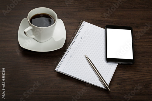 Top view on dark wooden office desk with notepad, pen, modern mobile with blank screen and cup of cofee.